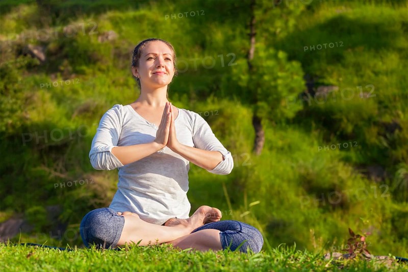 Meditation and relaxation yoga outdoors, young woman meditating and relaxing in Padmasana Lotus Pose with namaste Anjali mudra on green grass in forest