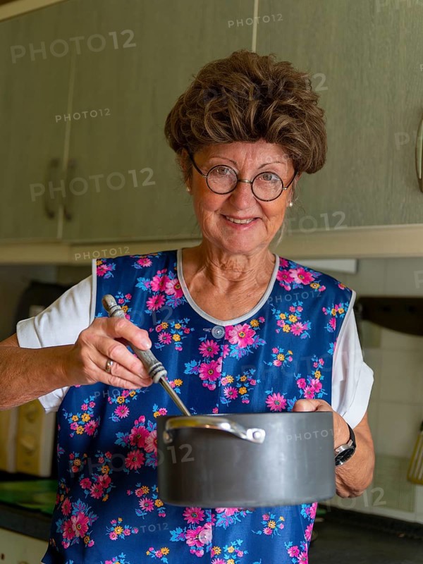 Granny with old smock apron, glasses and wig in the kitchen, holding cooking pot and ladle, Germany, Europe