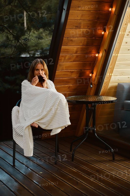 Cute young woman is sitting in an armchair on the terrace of a country house wrapped in a blanket with a cup of soothing tea and enjoying the fresh air