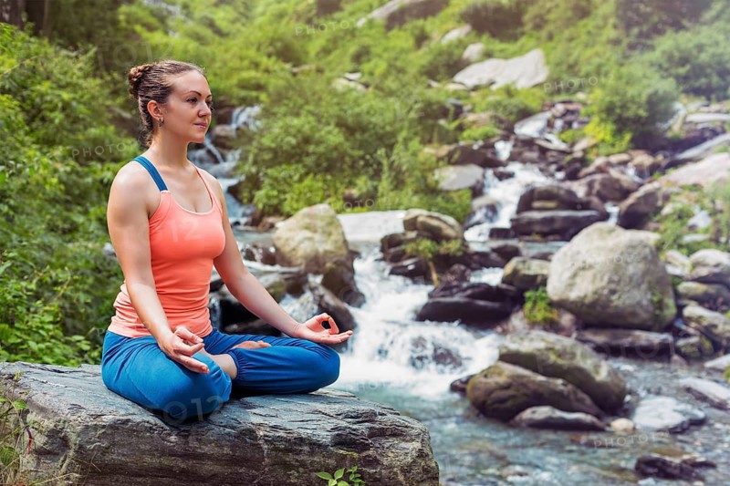 Woman in Hatha yoga asana Padmasana outdoors at tropical waterfall