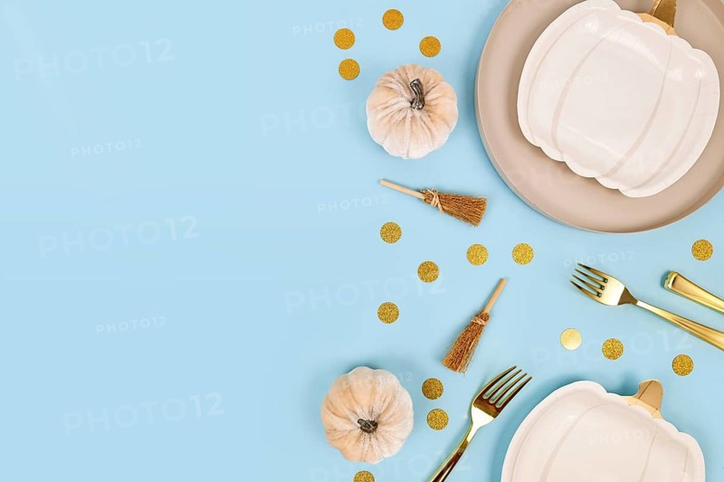 Halloween party flat lay with cream colored pumpkin shaped plates, witch brooms and golden confetti on blue background with copy space