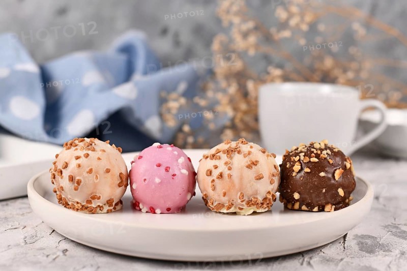 Small colorful cake balls glazed with white, pink and brown chocolate with sprinkles in a row on white plate