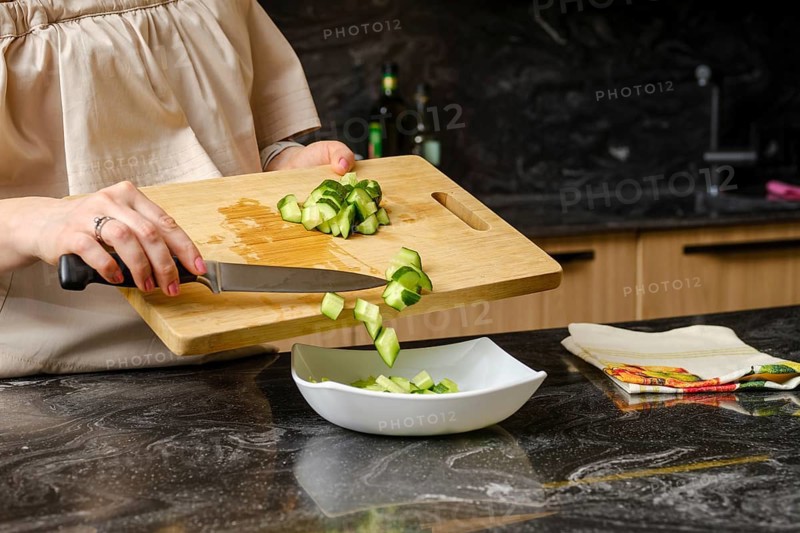 Closeup of hands of woman dropping a sliced cucumber into a dish with salad