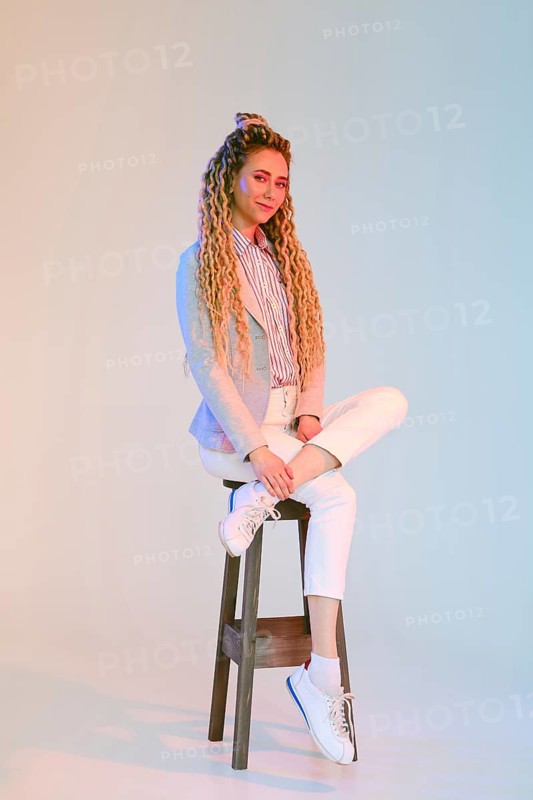 Young woman with dreadlocks in jeans and jacket sits on chair under colorful lights