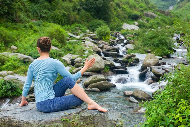 Yoga exercise outdoors, woman doing Ardha matsyendrasanaasana asana, half spinal twist pose at tropical waterfall in Himalayas in India