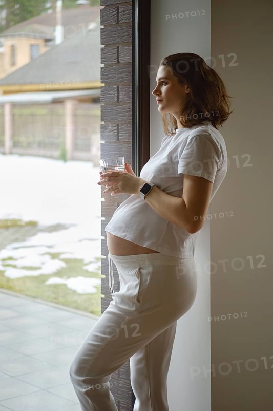 Lovely pregnant woman with glass of pure clean water in hand looks out of her kitchen window