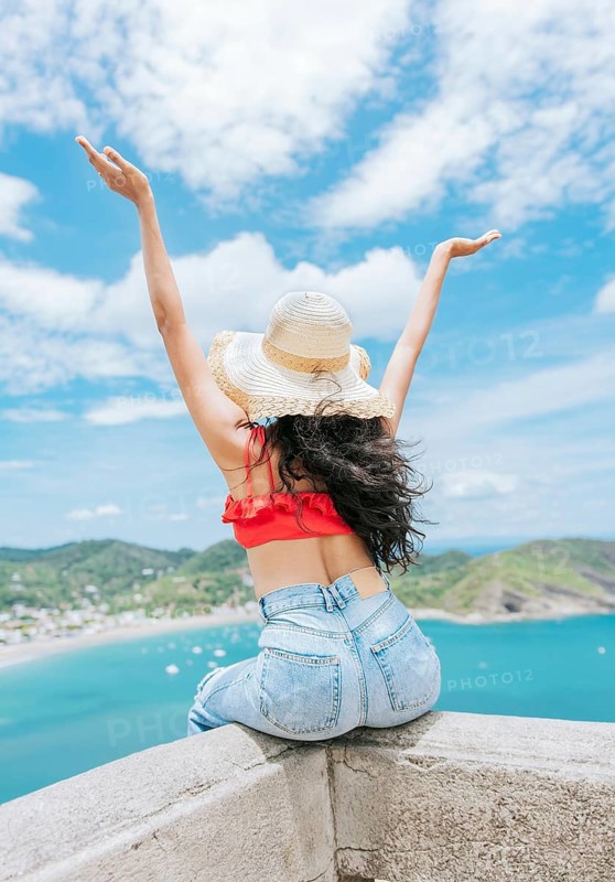 Lifestyle of woman on vacation sitting spreading hands to the beach. Tourist girl in hat spreading arms at a viewpoint looking at the beach