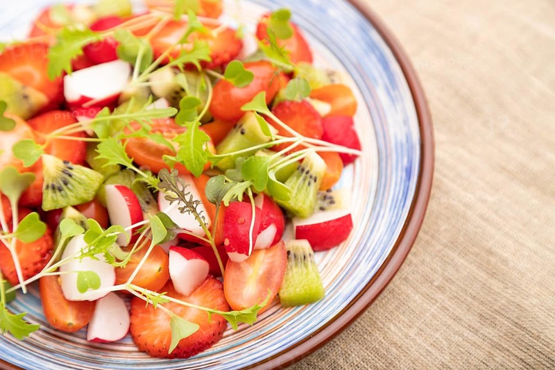 Vegetarian fruits and vegetables salad of strawberry, kiwi, tomatoes, microgreen sprouts on black concrete background and linen textile. Side view, close up, selective focus