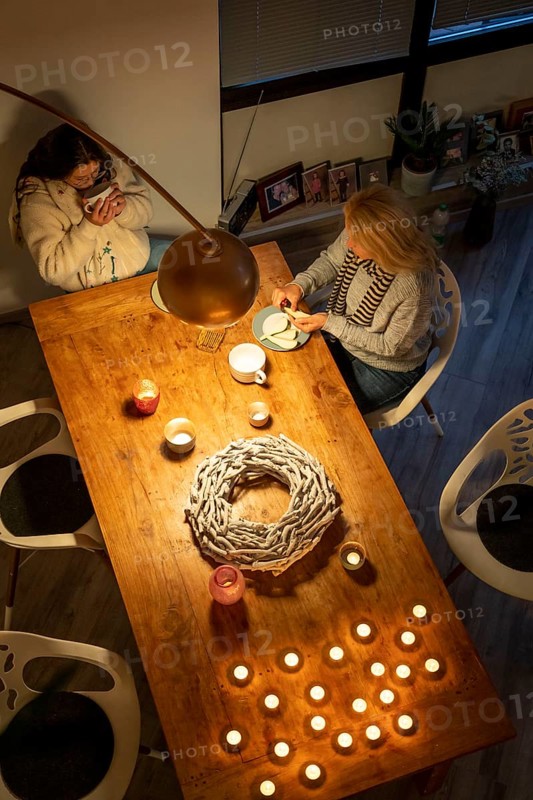 Symbolic image energy saving, cold winter, energy crisis, cold flat, mother and daughter dressed warmly at dinner, only one lamp on, candlelight, heating on to a minimum