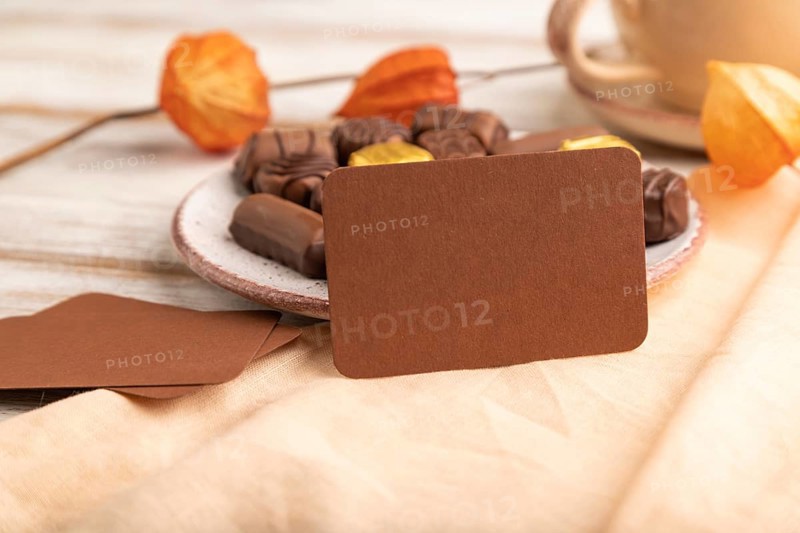 Brown business card with chocolate candies and physalis flowers on a white wooden background and orange textile. side view, close up, selective focus
