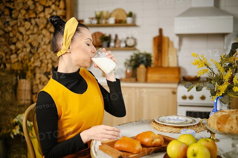 Woman drinking milk while sitting at the table in rustic style kitchen. The table is served with fresh buns, apples and decorated with wild flowers