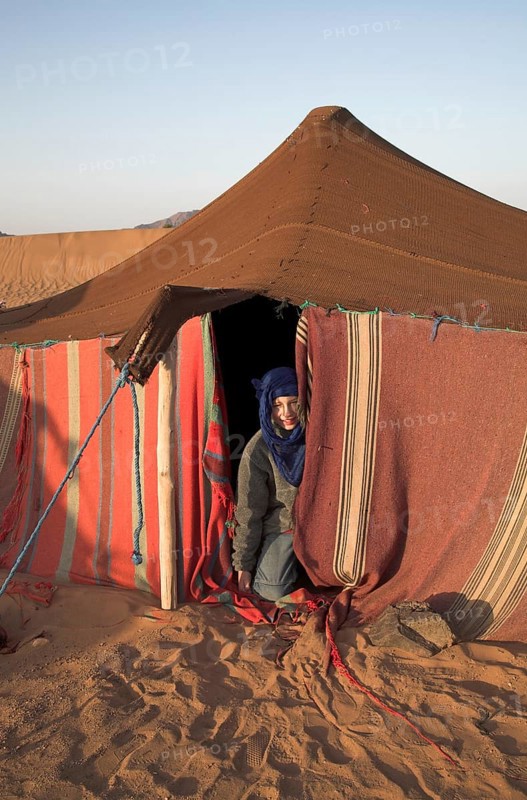 Model Released Young tourist boy dressed with blue nomad head scarf kneels at the entrance to a Berber tent, Sahara desert, near Zagora, Morocco, north Africa, Africa