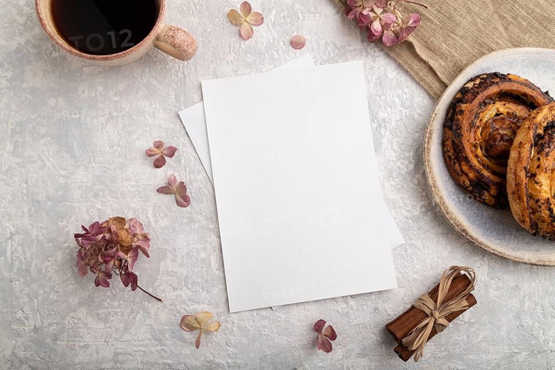 White paper invitation card, mockup with hydrangea flowers, bun, cup of coffee, linen textile, cinnamon on gray concrete background. Blank, flat lay, top view, still life, copy space