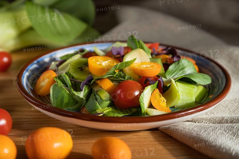 Vegetarian salad of pac choi cabbage, kiwi, tomatoes, kumquat, microgreen sprouts on a wooden background and linen textile. Hard light, contrast. Side view, close up, selective focus