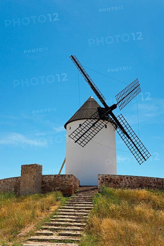 A historic windmill with black wings stands under a clear blue sky, surrounded by grassy hills and a stone staircase, Alcazar de San Juan, Ciudad Real, Castilla-La Mancha, Route of Don Quixote, Spain, Europe