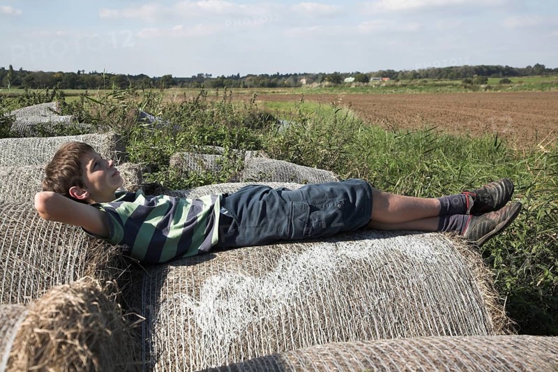 Model released image of young boy laying on bales of straw looking up at the sky Suffolk, England, UK