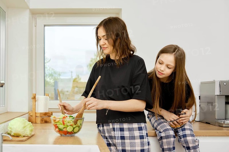The daughter looks out from behind her mother and watches her stir vegetables in a bowl