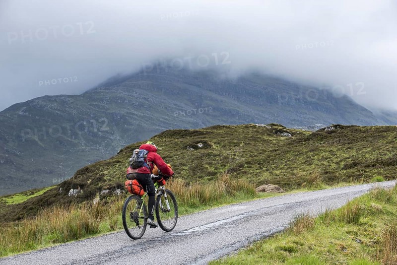 Lonely biker cycling through the Scottish Highlands on heavily laden touring bicycle along single track road in bad rainy weather, Scotland, UK