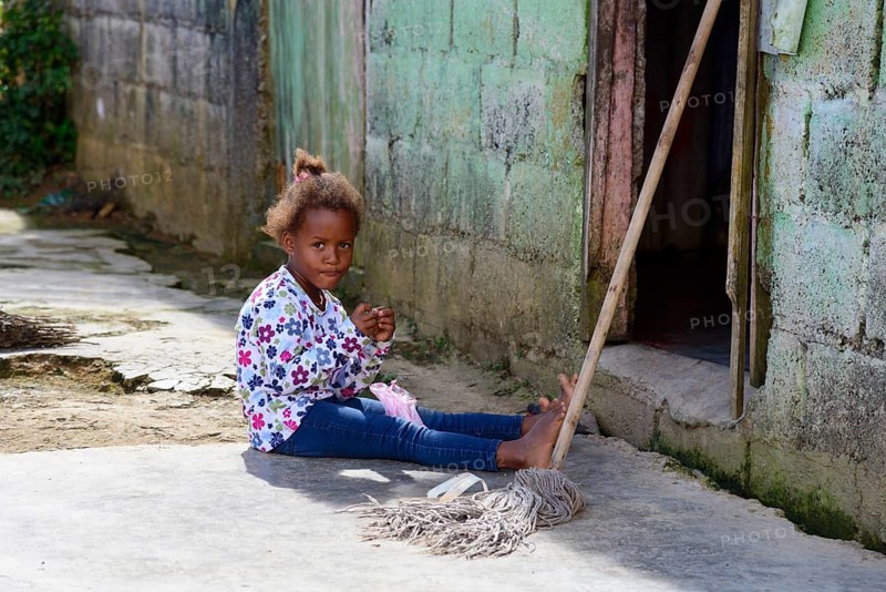A young girl plays on the ground next to a broom in front of a house in the countryside, between Pedro Sanchez and Miches, Cordillera Oriental, Dominican Republic, Hispaniola, Caribbean, America, Central America