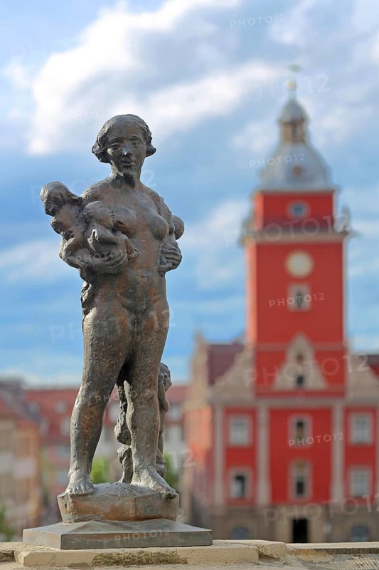 Sculpture Caritas by Bernd Göbel 2021 with blurred Old Town Hall, historical, allegory, Greek, mythology, love, virtue, woman standing with baby carrying, arms, depth of field, blur, horse trough, main market, Gotha, Thuringia, Germany, Europe