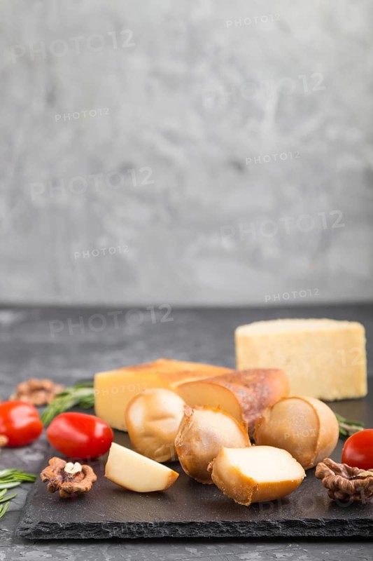 Smoked cheese and various types of cheese with rosemary and tomatoes on black slate board on a black concrete background. Side view, copy space, selective focus