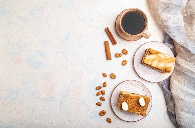 Honey cake with milk cream, caramel, almonds and a cup of coffee on a white concrete background and linen textile. Top view, flat lay, copy space