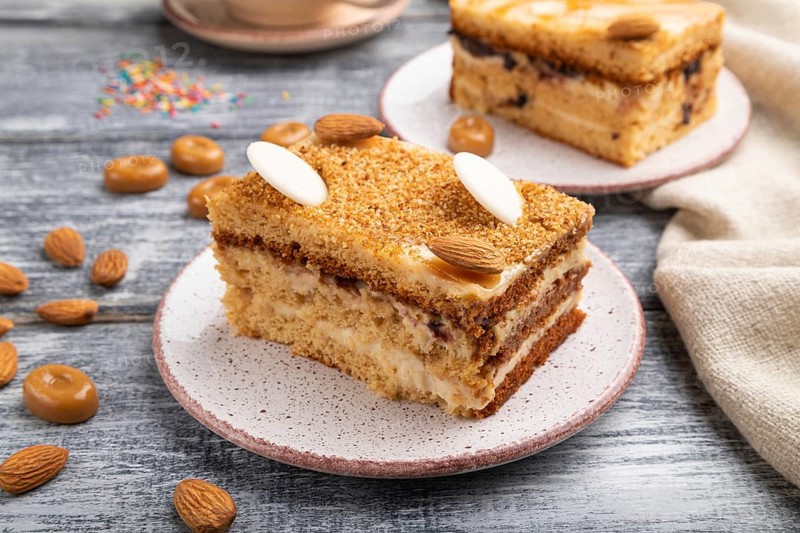 Honey cake with milk cream, caramel, almonds and a cup of coffee on a gray wooden background and linen textile. Side view, close up