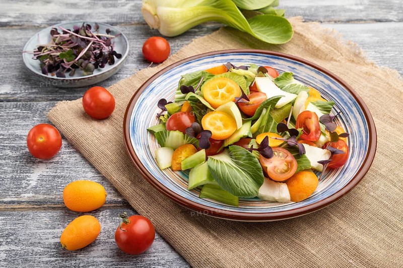 Vegetarian salad of pac choi cabbage, kiwi, tomatoes, kumquat, microgreen sprouts on gray wooden background and linen textile. Side view, close up
