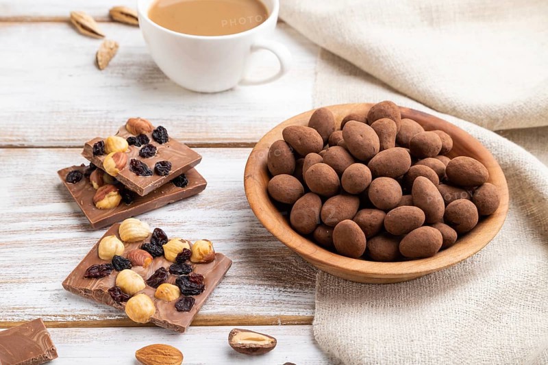Almond in chocolate dragees in wooden plate and a cup of coffee on white wooden background and linen textile. Side view, close up