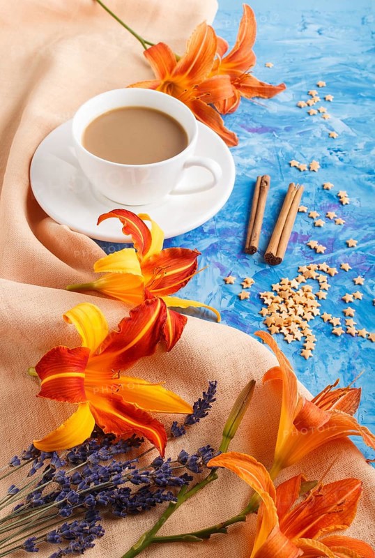 Orange day-lily and lavender flowers and a cup of coffee on a blue concrete background, with orange textile. Morninig, spring, fashion composition. side view