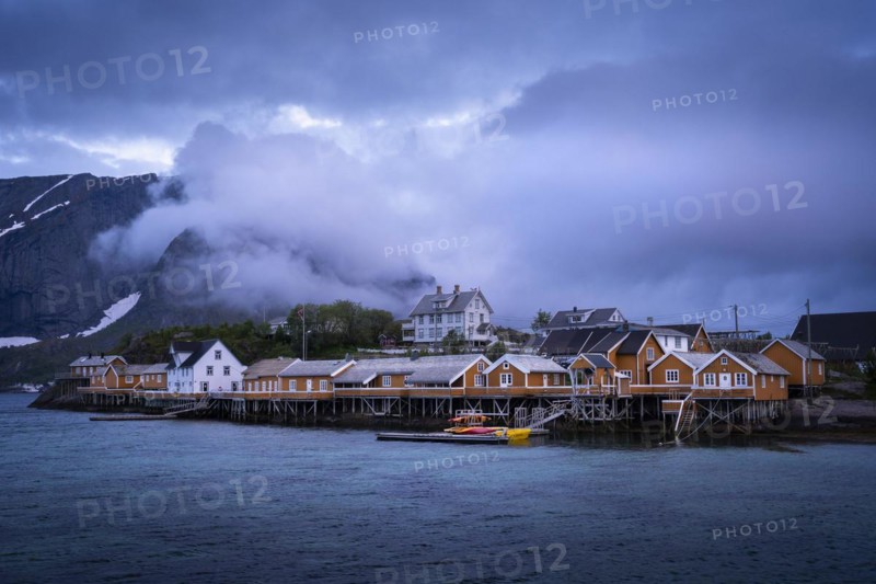 The village of Sakrisoy with its typical yellow or ochre-coloured wooden houses on wooden stilts (rorbuer) by the sea. At night at the time of the midnight sun. Clouds in the sky and in the mountains. Early summer. Sakrisoy, Moskenesoya, Lofoten, Norway, Europe