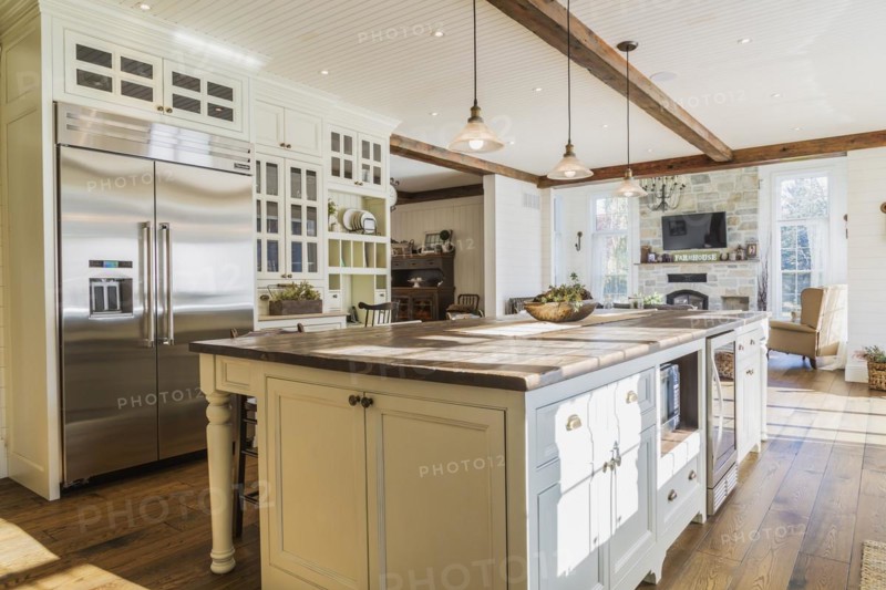 White painted maple wood antique style cabinets with tan coloured quartz countertops, island with brown stained wood plank countertop and high back bar stools in kitchen with stained oak wood floorboards inside country style home, Quebec, Canada, North America
