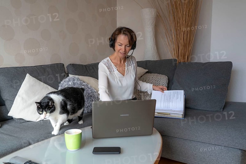 Woman, mid-50s, works from home, with laptop and communicates with colleagues via headset, home office, on the sofa