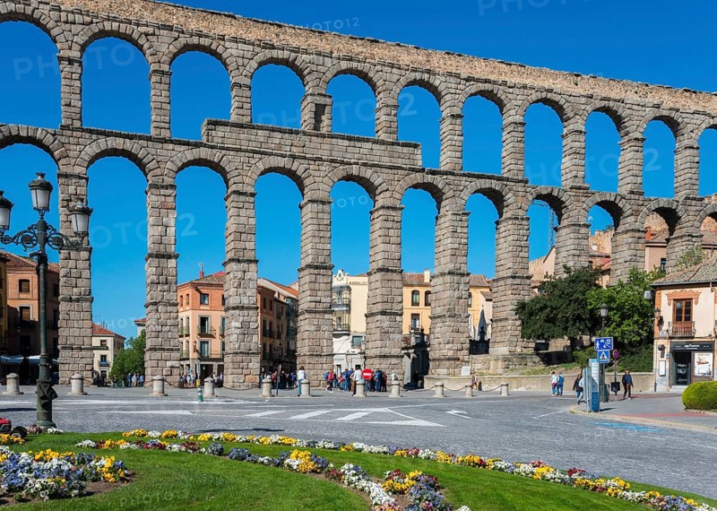 A historic aqueduct with stone arches spans a town with a flowerbed in the foreground under a blue sky, Aqueduct, Segovia, Castilla y León, Leon, Spain, Europe