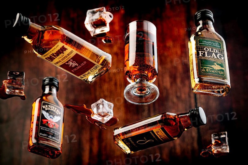 Four whisky bottles and a glass with spilt whisky and melted ice against a wooden background