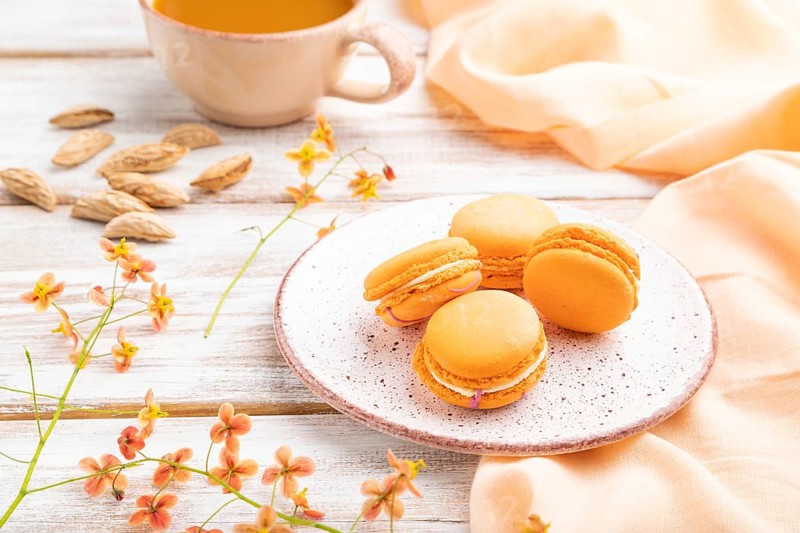 Orange macarons or macaroons cakes with cup of apricot juice on a white wooden background and orange linen textile. Side view, close up