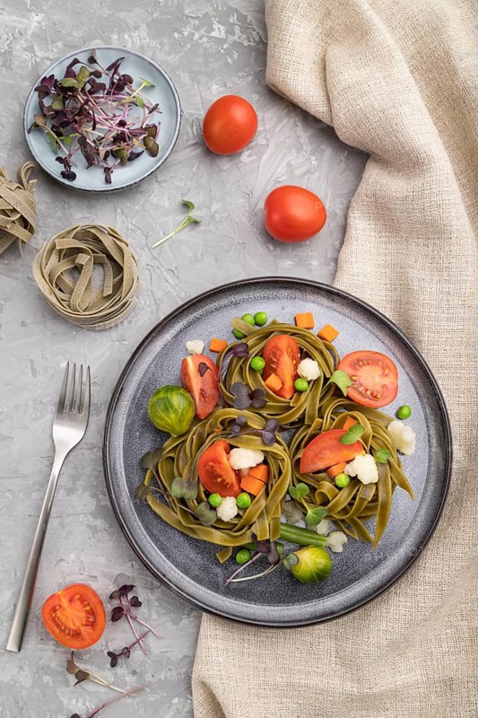 Tagliatelle green spinach pasta with tomato, pea and microgreen sprouts on a gray concrete background and linen textile. Top view, flat lay, close up