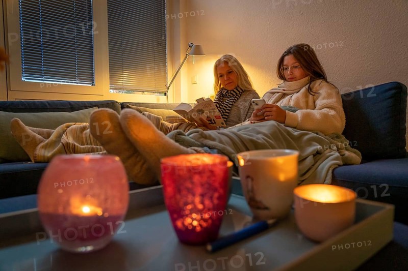 Symbolic image energy saving, cold winter, energy crisis, cold flat, mother and daughter lying on the sofa, warmly dressed, candlelight, heating on to a minimum