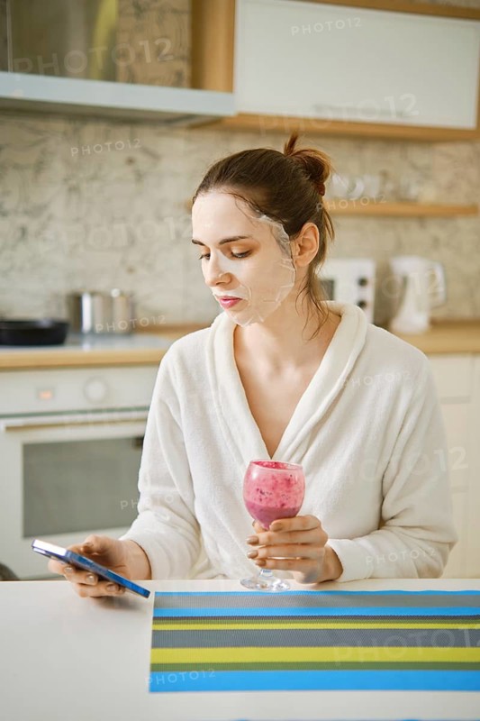 A woman applied a cosmetic mask to her face and spends time sitting in the kitchen with a smartphone and a berry smoothie