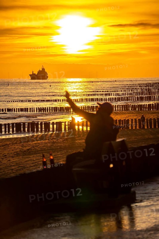 Sunset on the beach of Zoutelande, beach with wooden pile breakwaters, tourists, cargo ship sailing towards the Westerschelde, Zeeland, Netherlands