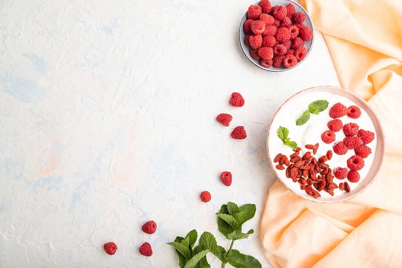 Yogurt with raspberry and goji berries in ceramic bowl on white concrete background and orange linen textile. top view, flat lay, copy space