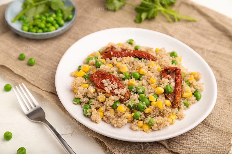 Quinoa porridge with green pea, corn and dried tomatoes on ceramic plate on a white wooden background and linen textile. Side view, close up, selective focus