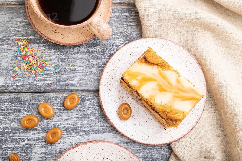 Honey cake with milk cream, caramel, almonds and a cup of coffee on a gray wooden background and linen textile. Top view, flat lay, close up