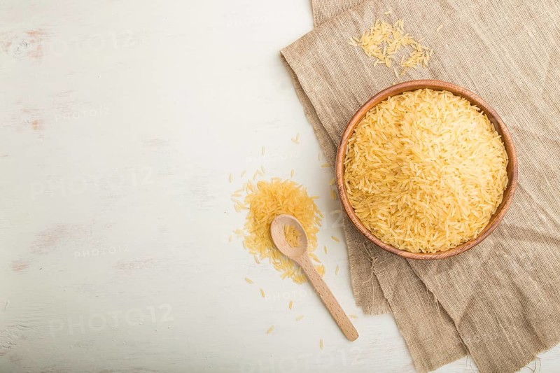 Wooden bowl with raw golden rice and wooden spoon on a white wooden background and linen textile. Top view, flat lay, copy space