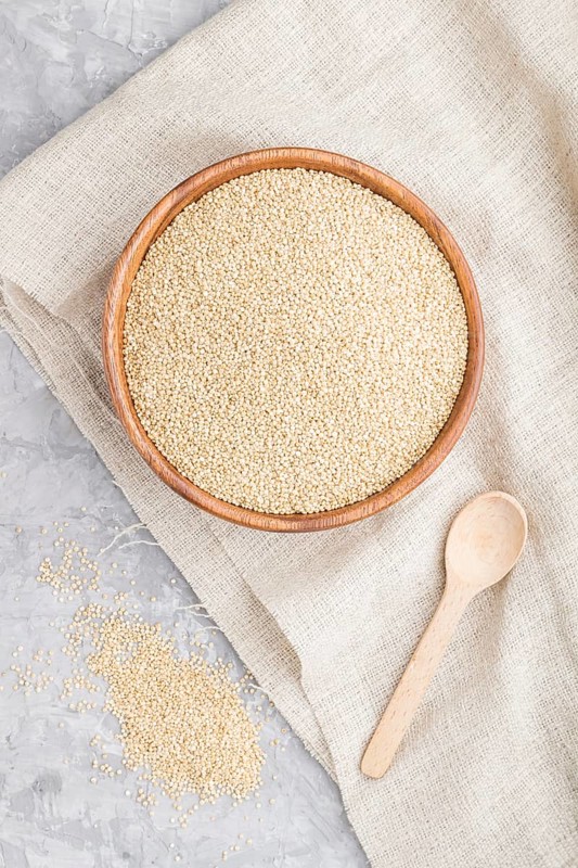 Wooden bowl with raw white quinoa seeds and wooden spoon on a gray concrete background and linen textile. Top view, flat lay, close up