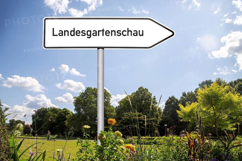 Symbolic image of the State Garden Show: Directional arrow with the inscription Landesgartenschau in a park (Compsosing)