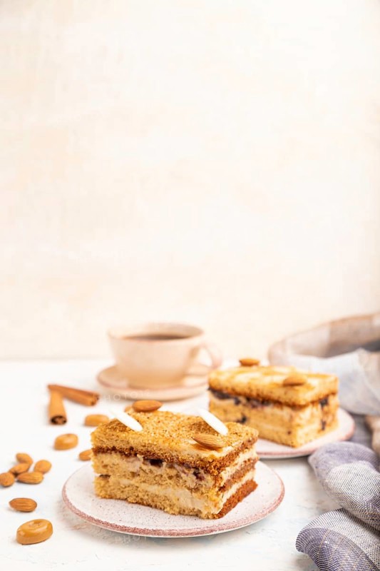 Honey cake with milk cream, caramel, almonds and a cup of coffee on a white concrete background and linen textile. Side view, copy space, selective focus