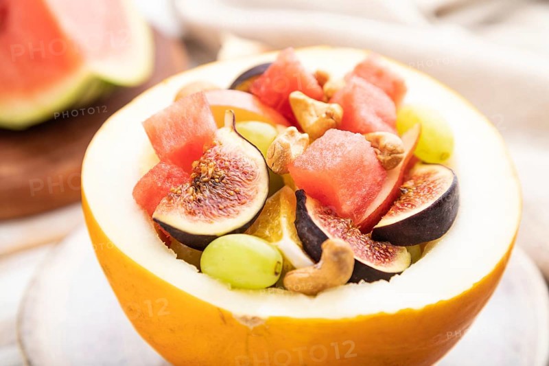 Vegetarian fruit salad of watermelon, grapes, figs, pear, orange, cashew on white wooden background and linen textile. Side view, close up, selective focus