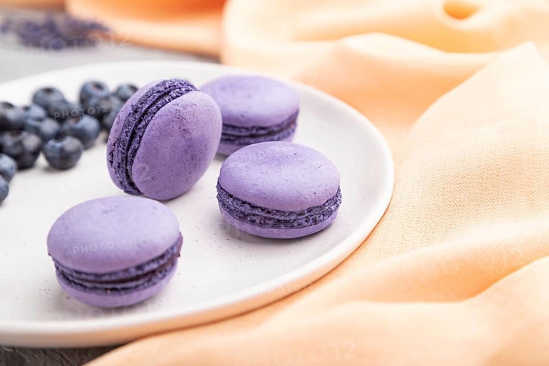 Purple macarons or macaroons cakes with blueberries on white ceramic plate on a gray concrete background and orange textile. Side view, close up, selective focus