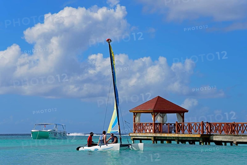 A colourful catamaran sails next to a jetty near a pavilion on the clear sea under a blue sky, Dominicus beach, Bayahibe, Dominican Republic, Hispaniola, Caribbean, America, Central America